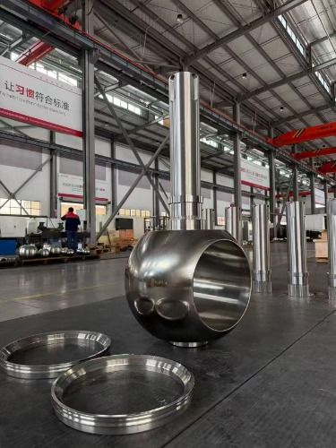 A large polished metal valve component and cylindrical parts are displayed in the company's industrial production workshop.